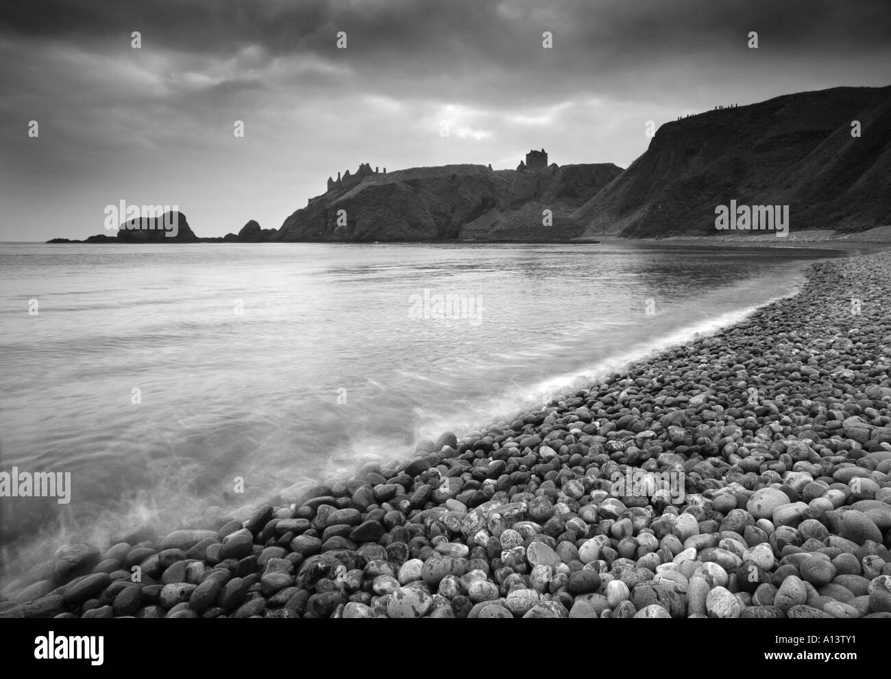 Black and white image of Dunnottar Castle, Stonehaven with pebbled wave lapped shore leading in to the castle with moody sky. Stock Photo