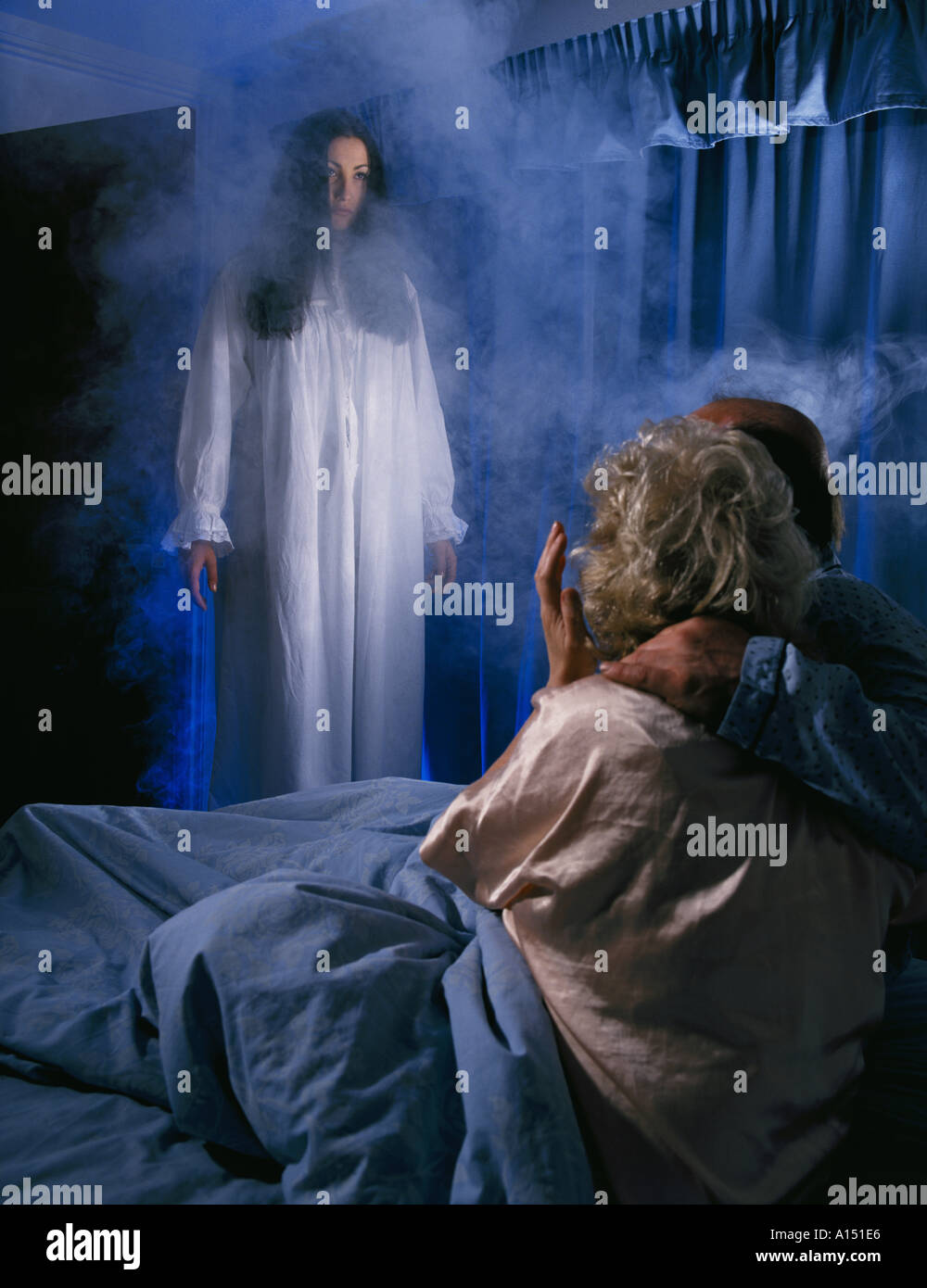 COUPLE IN BED HUGGING EACHOTHER WITH FEMALE GHOST FIGURE IN WHITE NIGHTY STANDING OVER THEIR BED MODEL RELEASED  SPOOKY SCARY SCARE FRIGHTENING SUPERNATURAL INTRUDER SPIRIT DEATH EXPERIENCE LIFE AFTERLIFE OUT OF BODY SMOKE MIST FOG MOODY RELATIONSHIP ELDERLY YOUTH Stock Photo