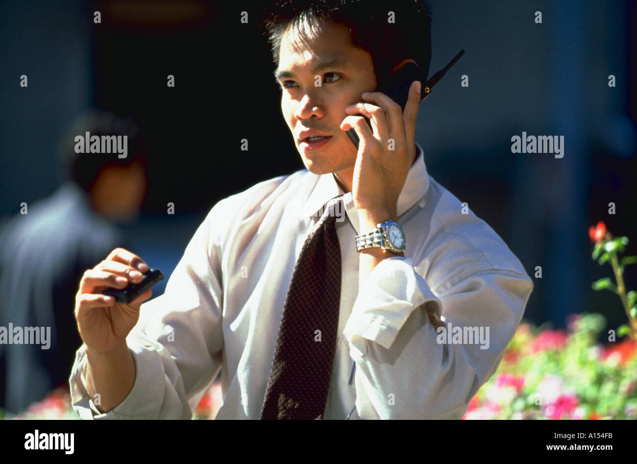 A young Asian businessman sits outside and talks on his cellular phone as he checks his pager Stock Photo