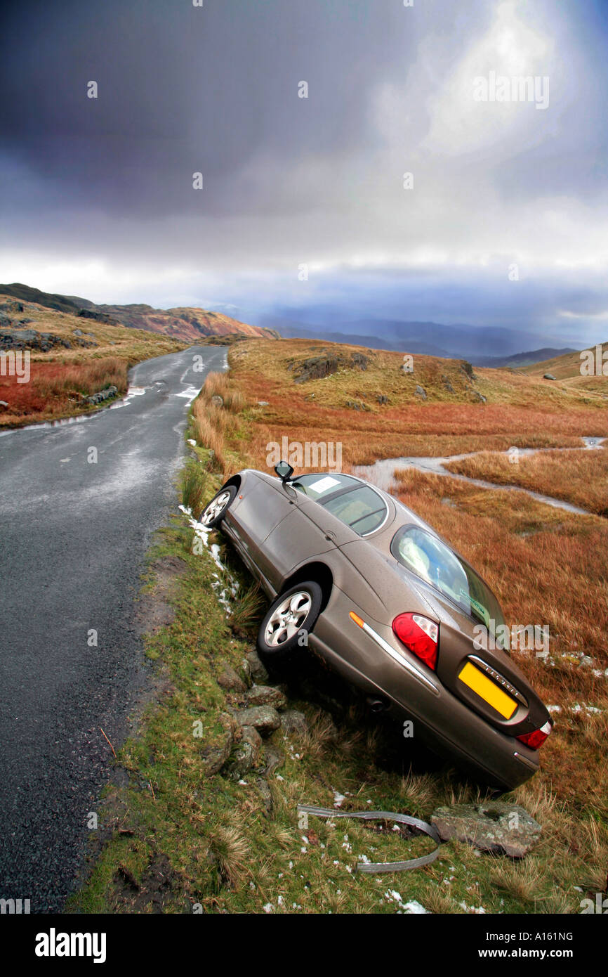 An SType Jaguar motor car lies in a ditch next to a wet mountain road