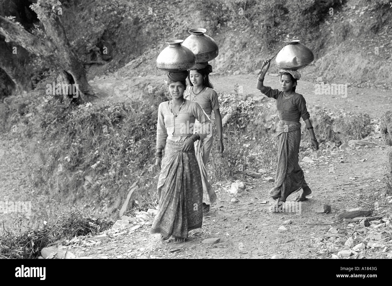 B/W of three Garwhali tribal women carrying metal pots full of water on their heads back to their village from the water source. Garhwal Himal, India Stock Photo