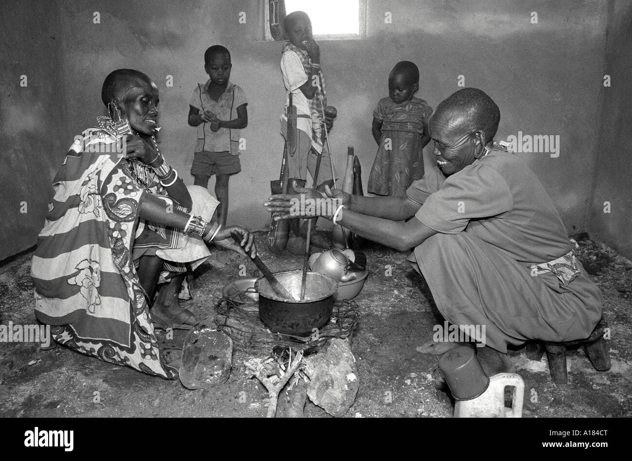 B/W of Maasai women with children, in traditional and modern dress, cooking over an open fire on the floor of their new house Nr. Kajiado, Kenya Stock Photo