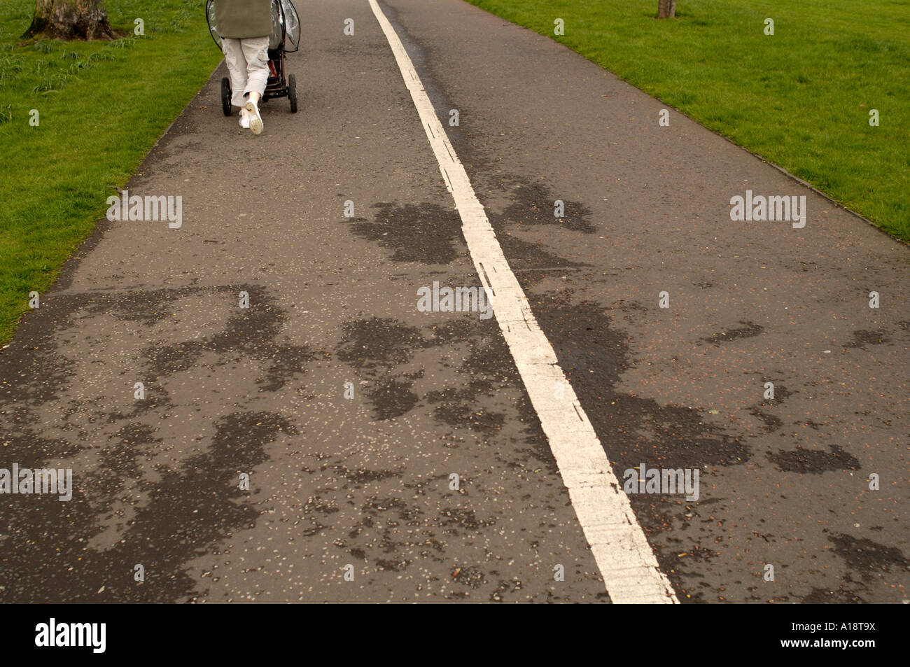 A woman pushing a childs pushchair along a grey path with white line and green grass Stock Photo