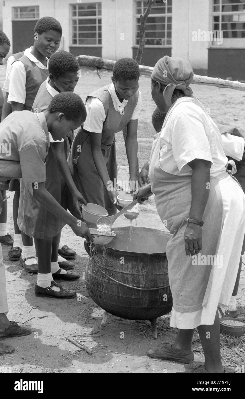 B/W of a cook serving schoolgirls their lunch from a large pot, their free meal is funded by a UK aid agency. Eswatini (Swaziland) Stock Photo