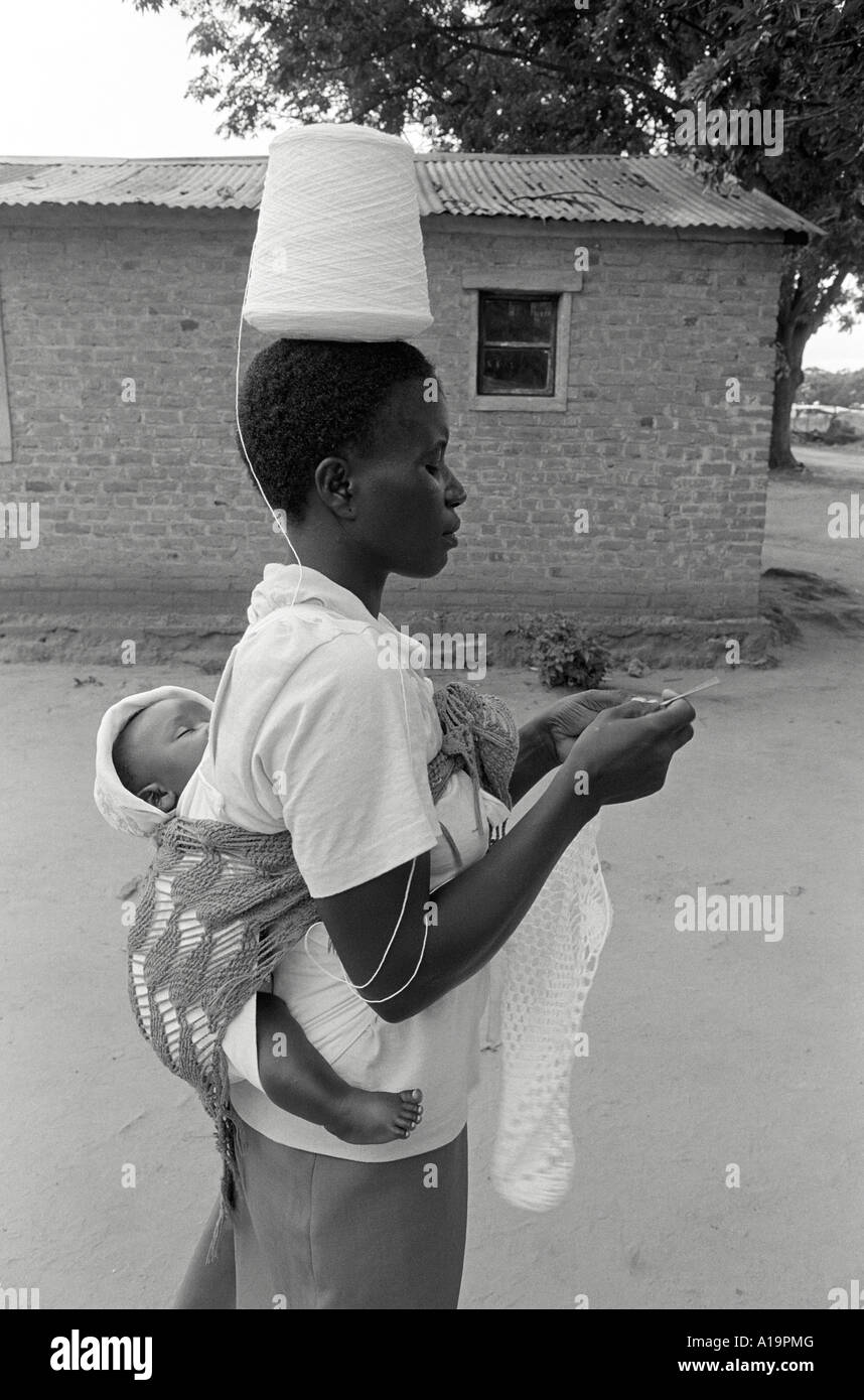 B/W of a farm worker with baby on her back, walking and crocheting  while balancing a spool of wool on her head.  Centenary, Zimbabwe Stock Photo