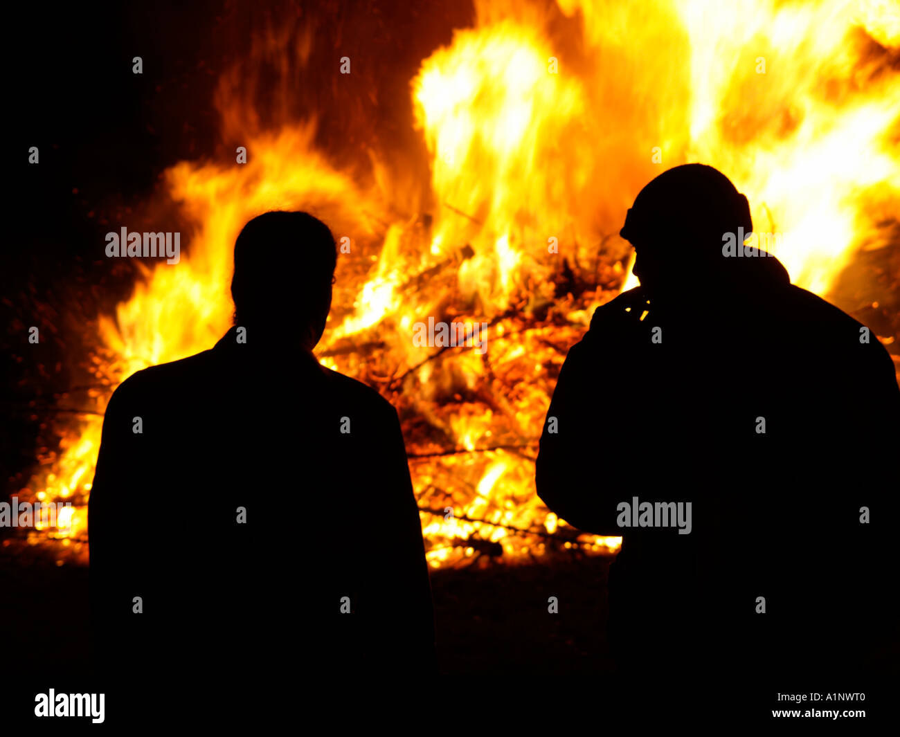 two men silhouetted in front of a huge fire bonfire from burning old christmas trees Stock Photo