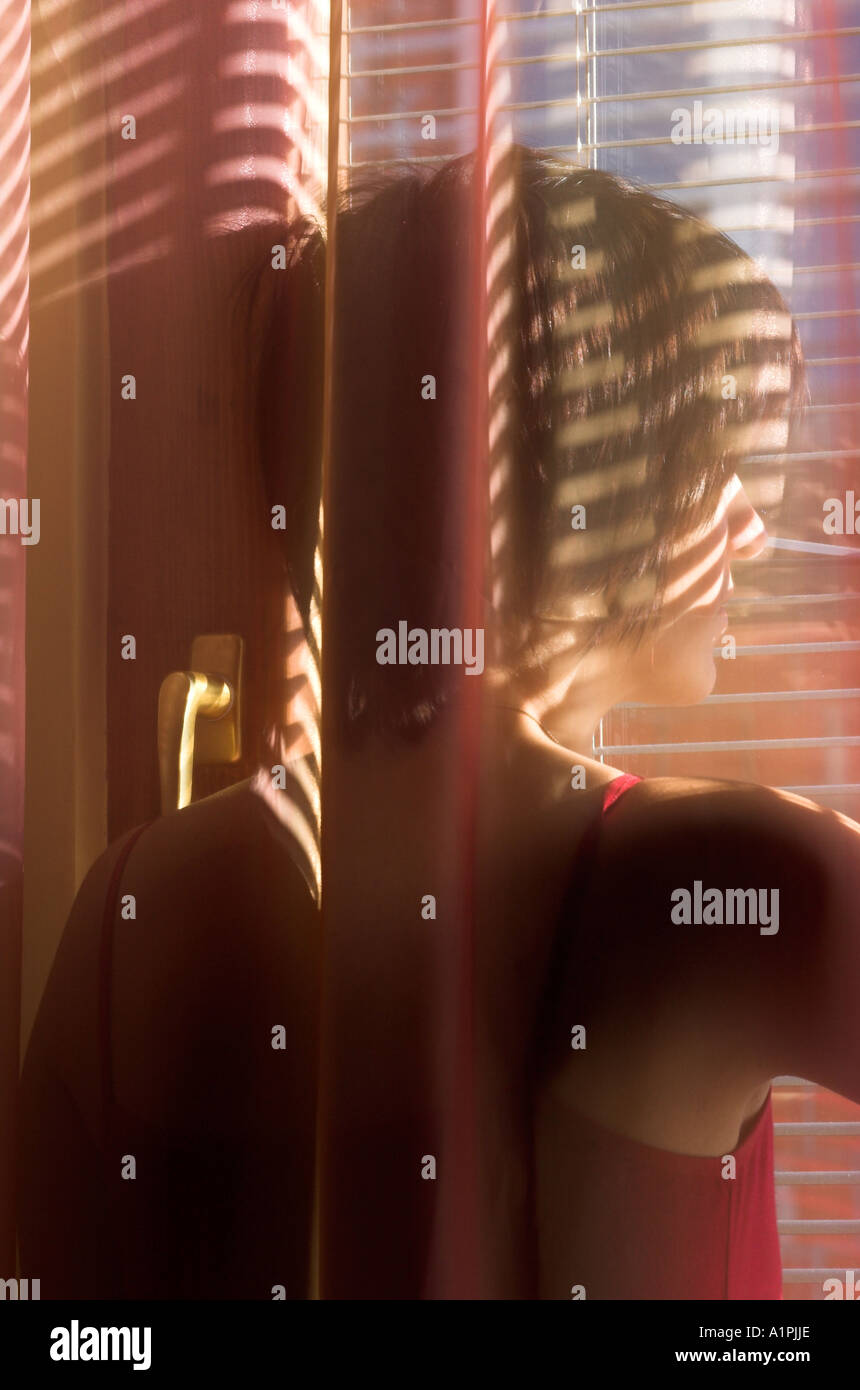 Young woman standing behind curtain looking through window blinds Stock Photo