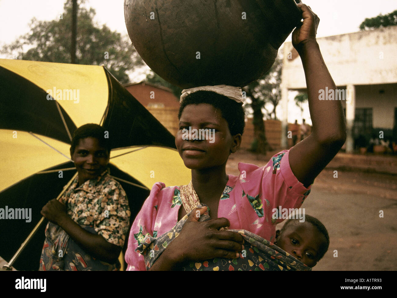 WOMAN CARRYING LARGE ROUND POT ON HER HEAD BABY ON BACK GOING TO MARKET DEC 1993 Stock Photo