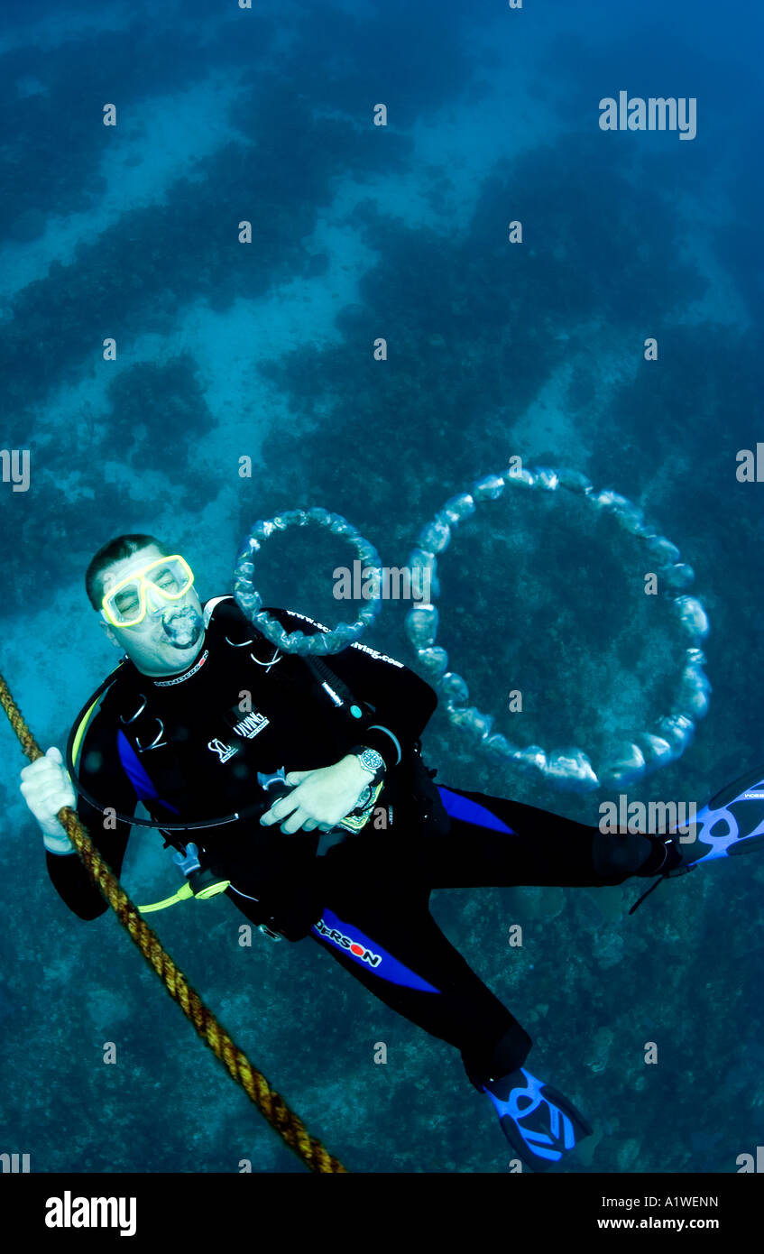 A MALE SCUBA DIVER BLOWS BUBBLE RINGS UNDERWATER WHILE HANGING ON AN