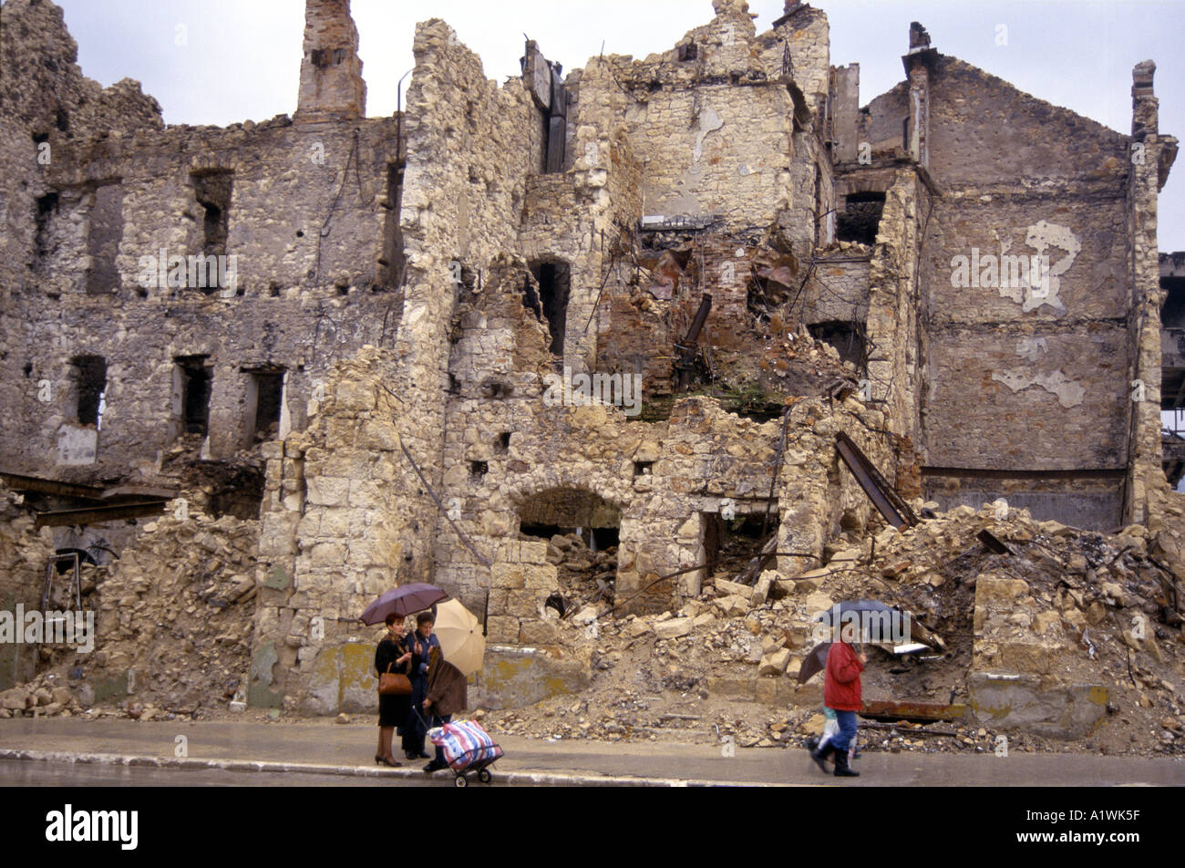 THREE WOMEN STANDING HOLDING UMBRELLAS TALKING ON THE STREET BESIDE THE REMAINS OF SEVERAL DESTROYED BUILDINGS IN MOSTAR 1994 Stock Photo