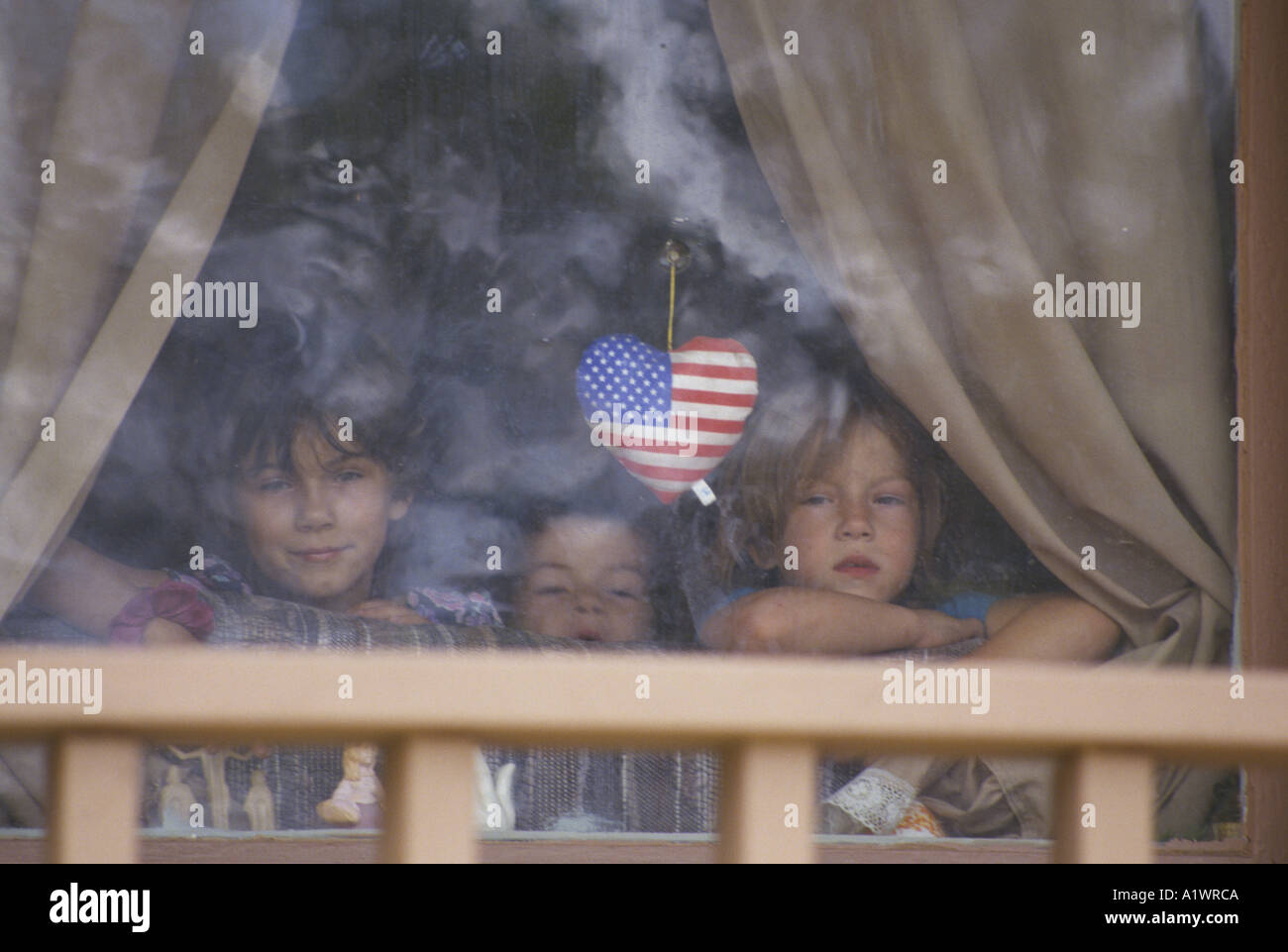 Three children looking out of window with stars and stripes shaped heart in a poor neighbourhood Stock Photo