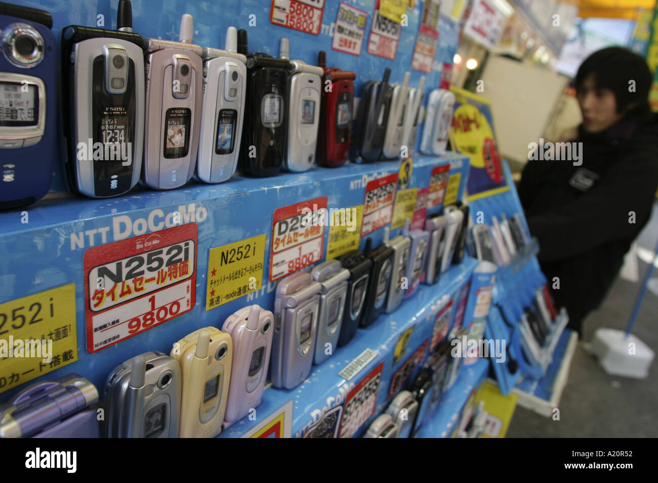 Teenager behind rows of mobile phones for sale on a NTT DoCoMo stand in an electronics store, Tokyo, Japan Stock Photo