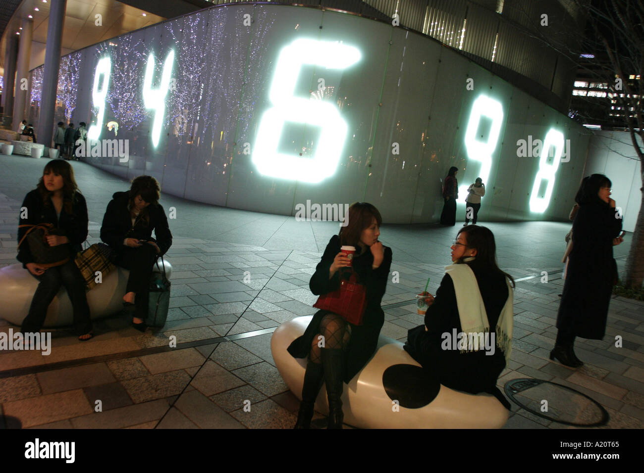 At night pedestrians sit beside a large digital number display on the wall of the TV Asahi television building in Roppongi Hills Stock Photo
