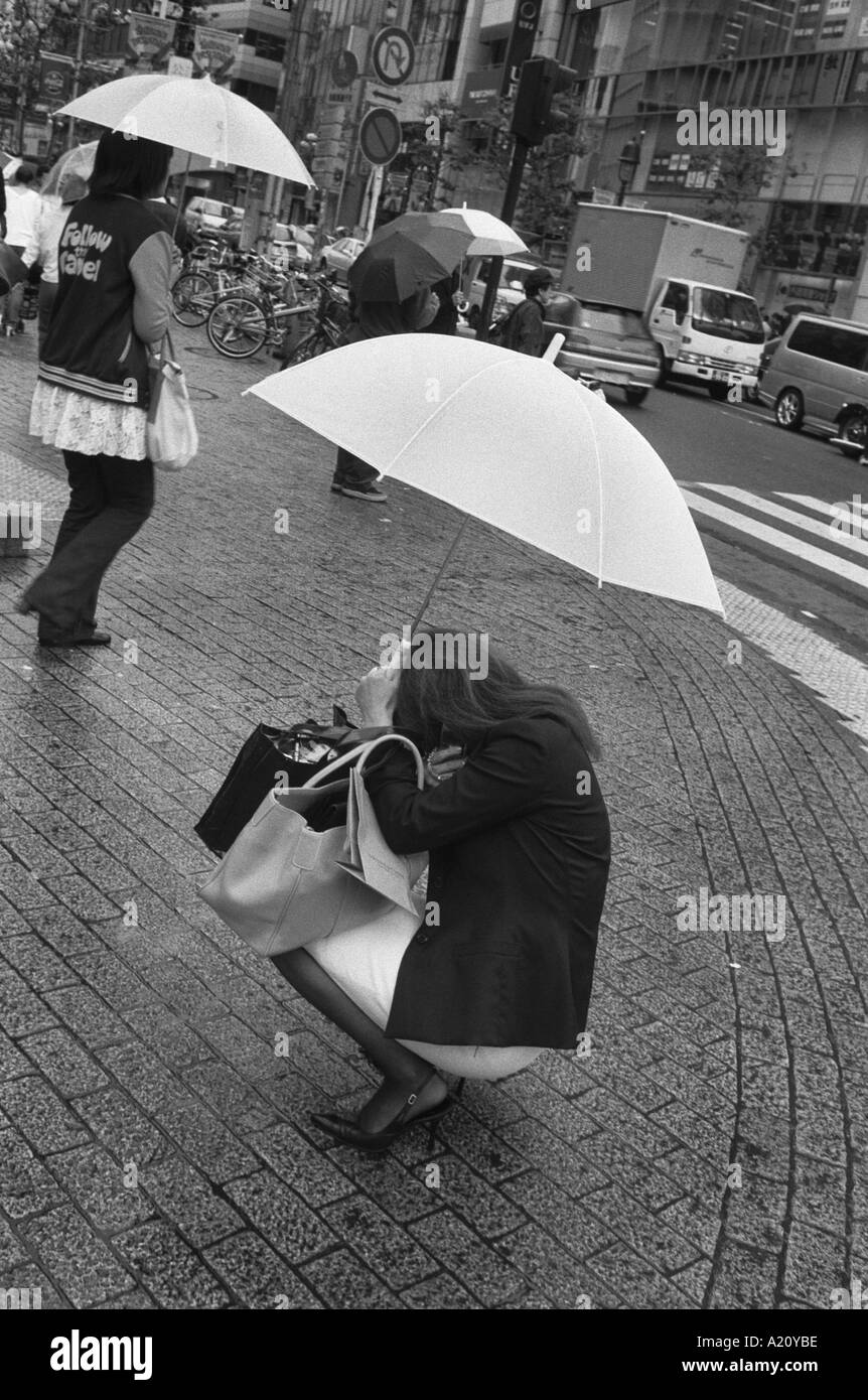 A woman squats in a busy Shibuya street under an umbrella to have a conversation on her mobile telephone, Tokyo, Japan Stock Photo
