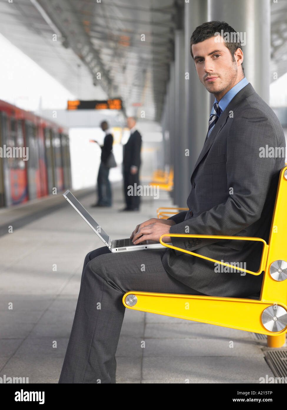 Businessman sitting on bench, working on laptop at Train Station, side view Stock Photo