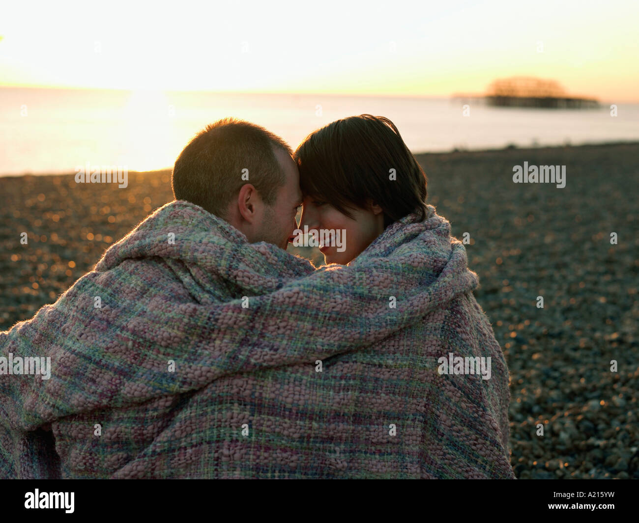 Couple wrapped in blanket sitting on beach at sunset Stock Photo - Alamy