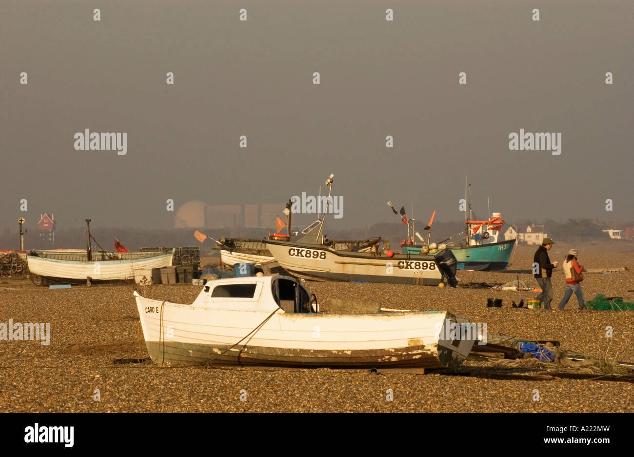 People walking by small fishing boats on beach at Aldeburgh. Sizewell B Nuclear Power Station is on horizon left of centre Stock Photo