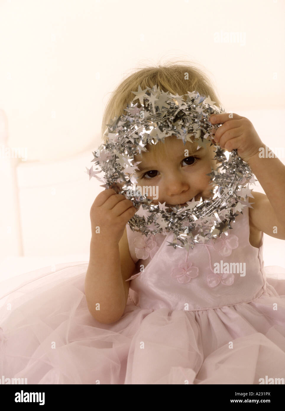 Toddler wearing a pink party dress peeking through a tinsel halo Stock Photo