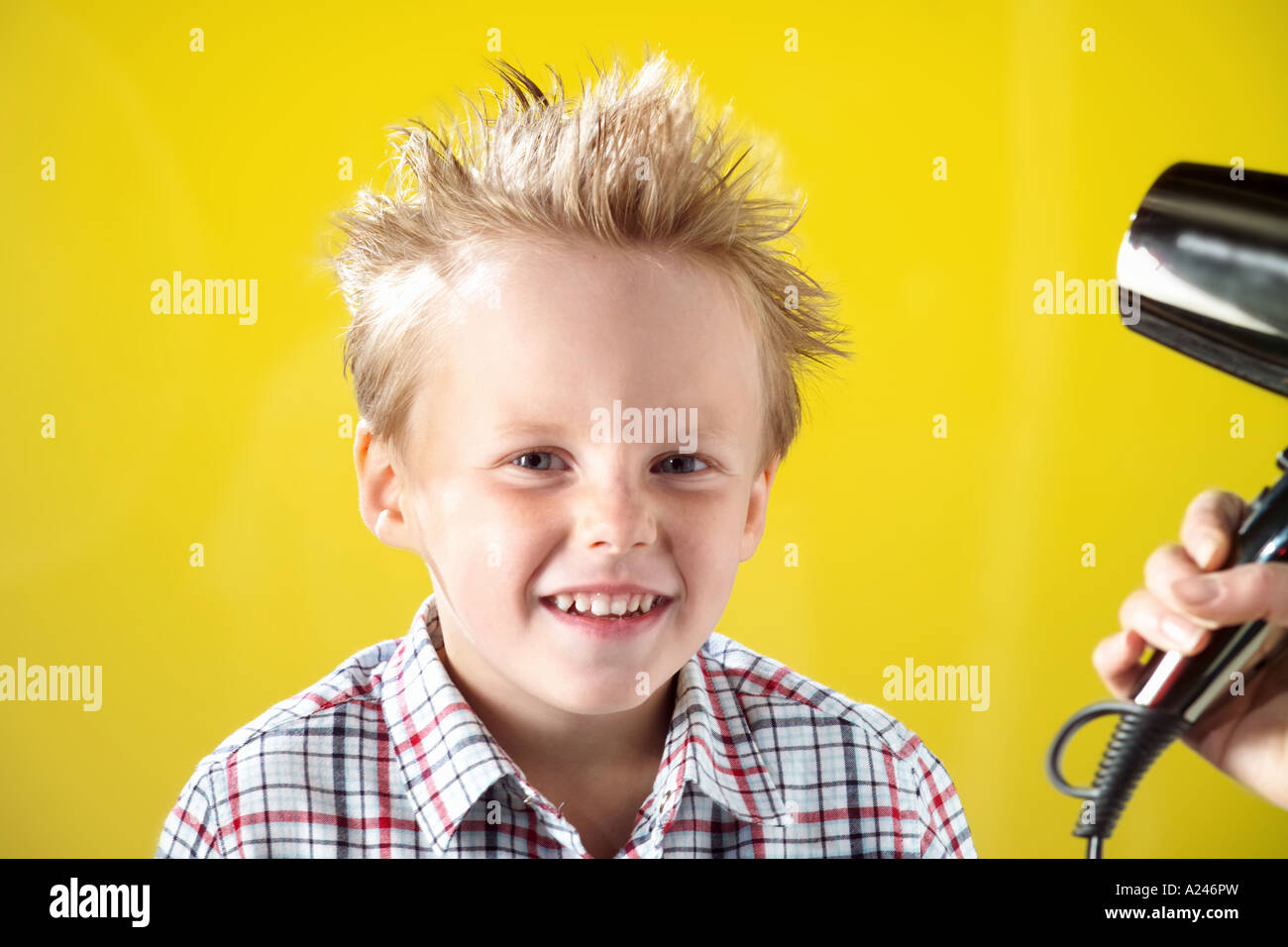 Blonde Boy aged 5-7 years against yellow studio background pulling funny faces while having his hair blow dried. Stock Photo