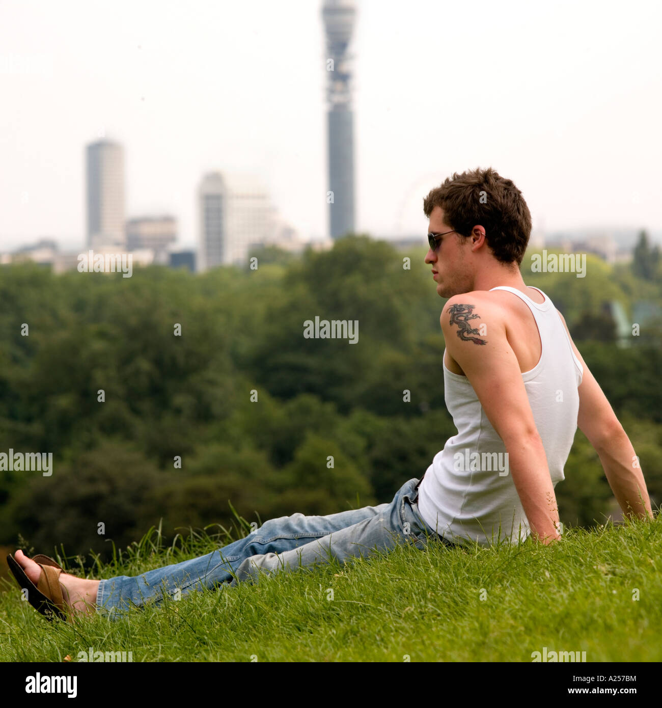 Young Man sits on Primrose Hill with the London skyline in the background in the summer Stock Photo