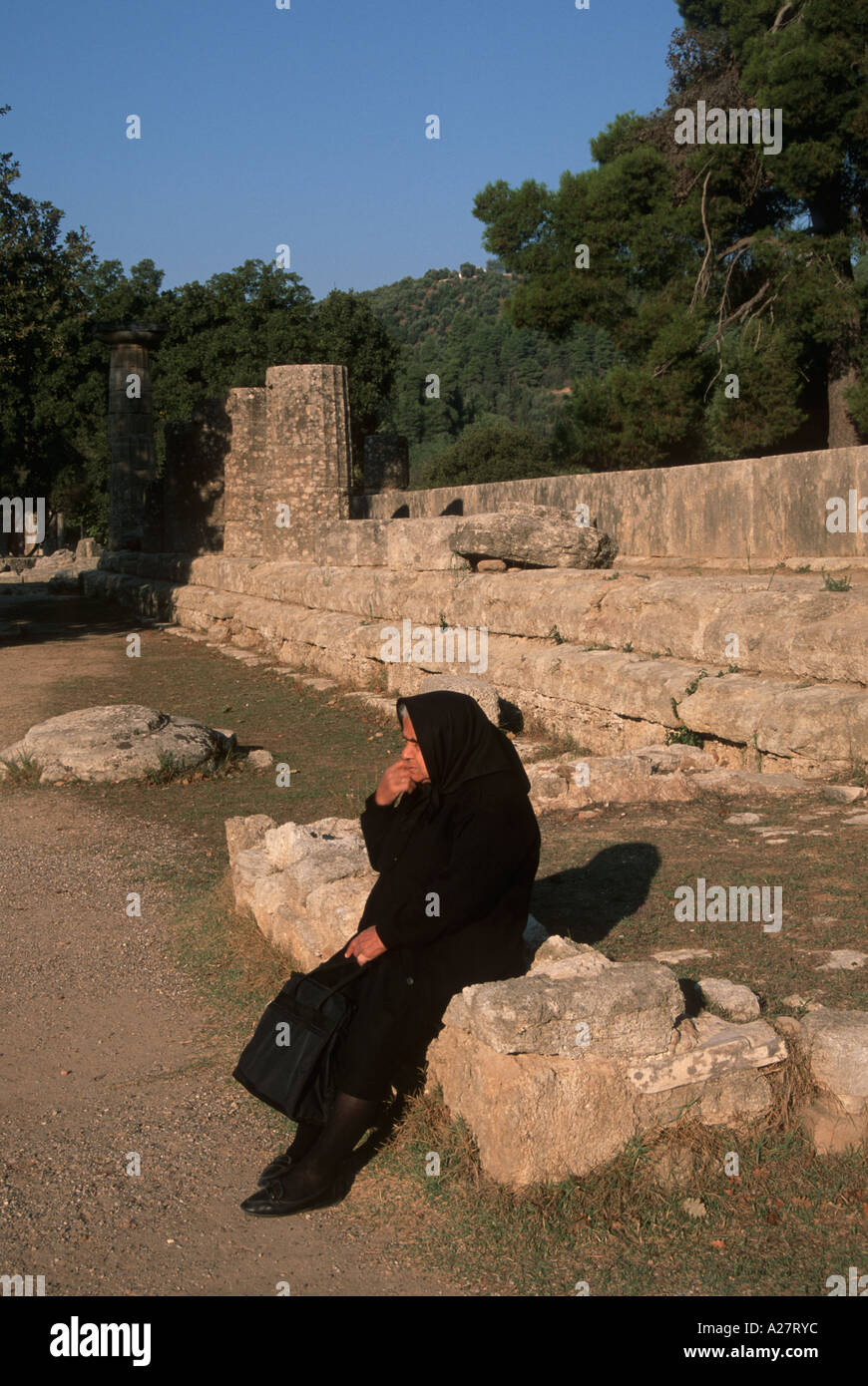 old woman with black dress seated on a ruin of Olympia Greece Stock Photo