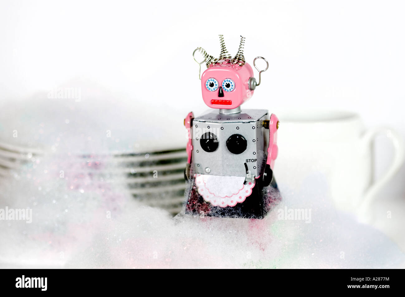 Female tin toy robot standing in front of dishes and covered in soap suds or washing up liquid bubbles Stock Photo