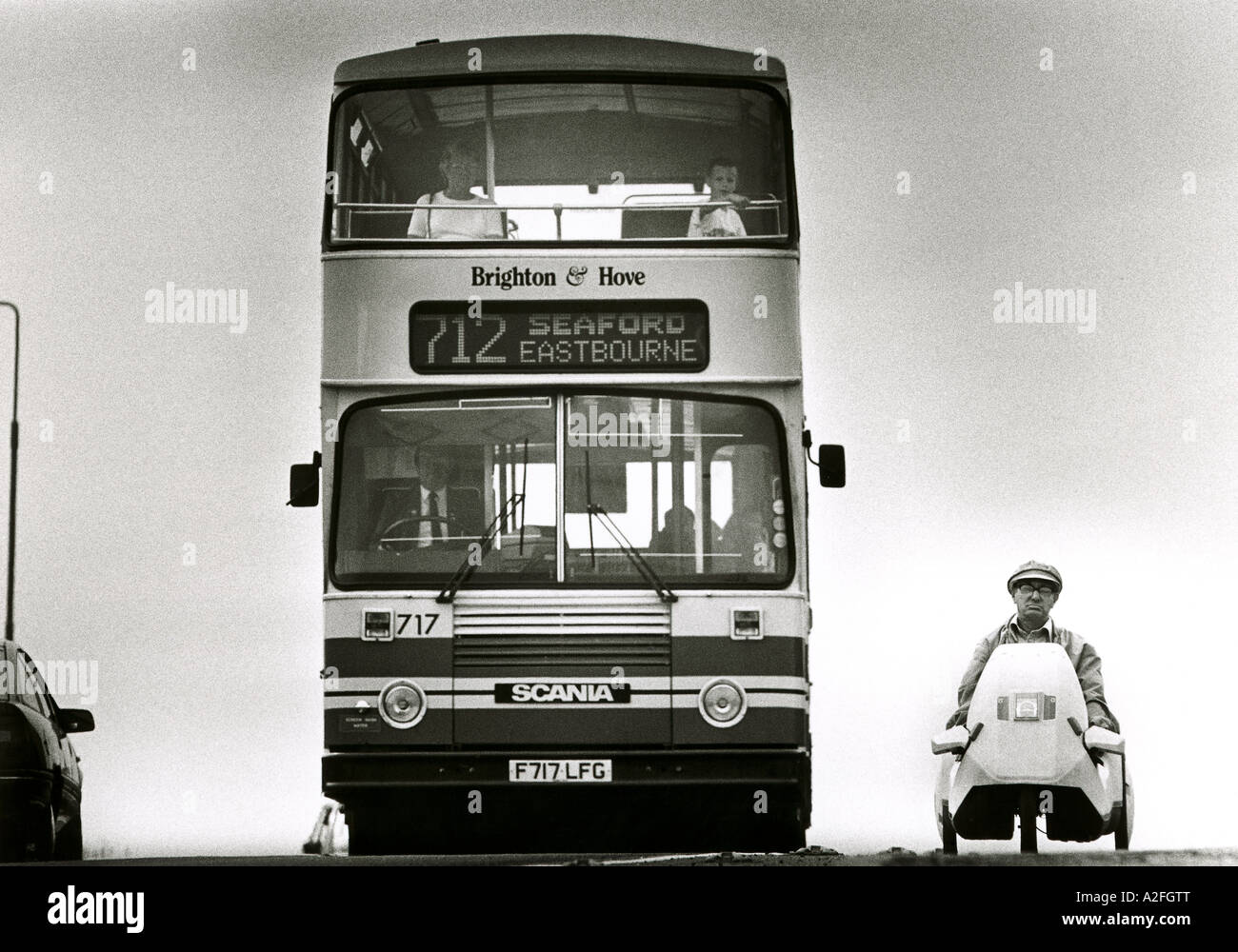 A grumpy faced man in a Sinclair C5  electric car being overtaken by a bus. Stock Photo