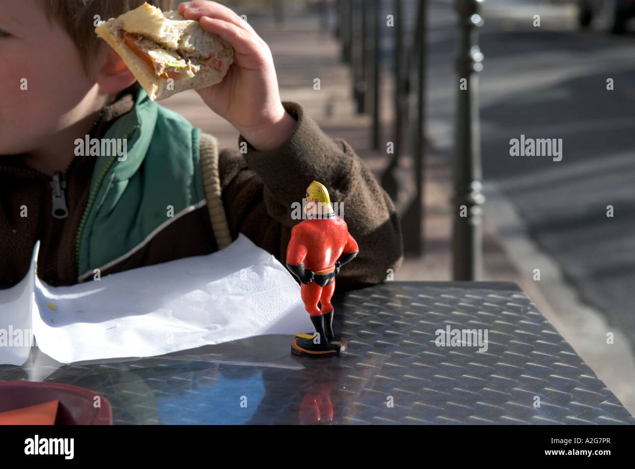 335 a small toy model of mr incredible stands on a cafe table looking at a young boy eating his sandwich Stock Photo