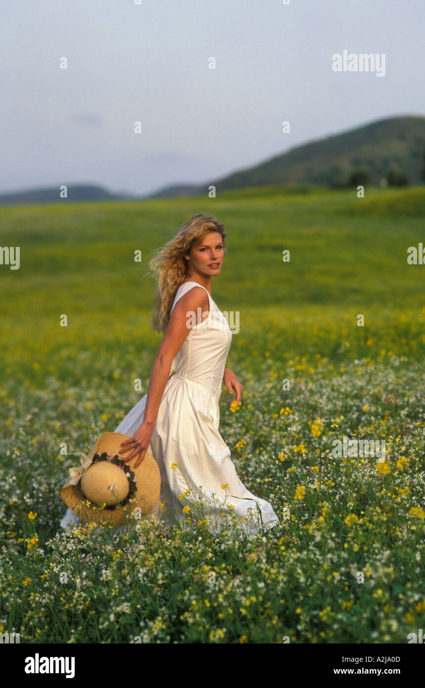 Blonde woman wearing a white sun dress and carrying a straw hat walks ...