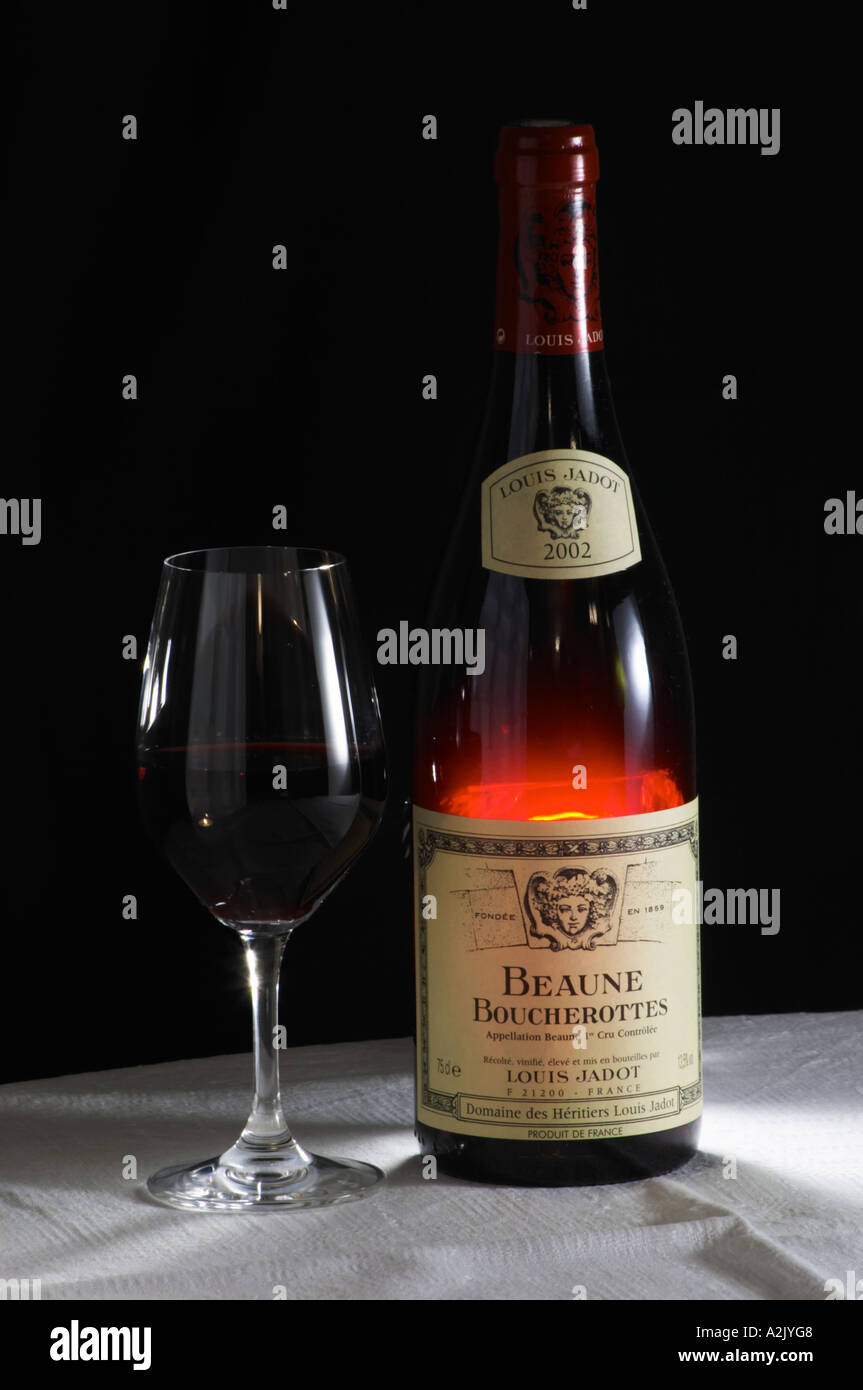 A bottle of Maison Louis Jadot Beaune Boucherottes 2002 red burgundy wine and a glass of red wine standing on a table top with a white cloth. Backlit backlight back light lit Black background, Maison Louis Jadot, Beaune Côte Cote d Or Bourgogne Burgundy Burgundian France French Europe European Stock Photo