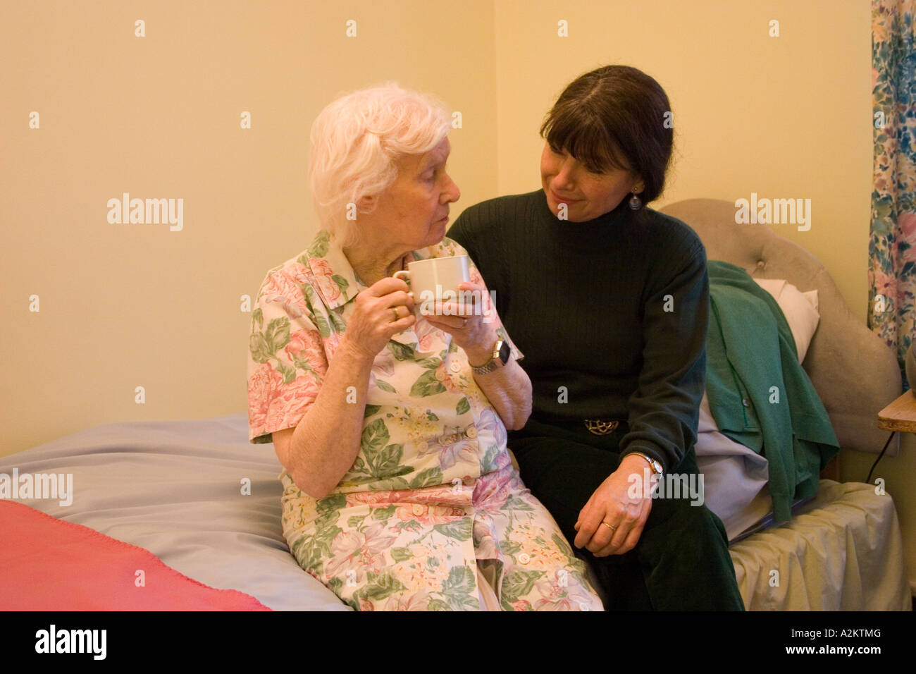 woman/daughter/care worker sitting on edge of bed with old lady/mother in her room/nursing home Stock Photo