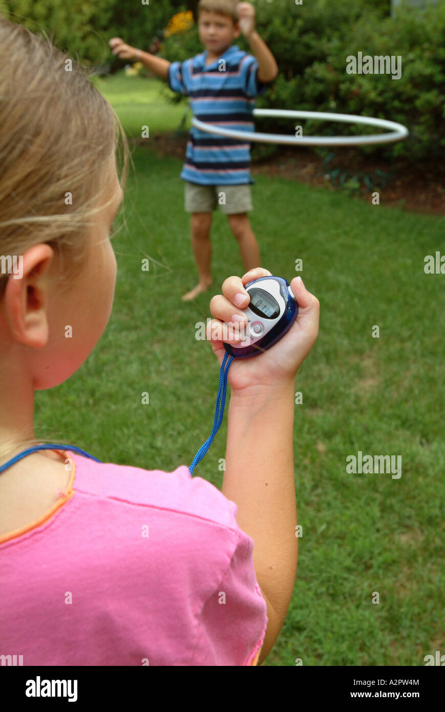 10 year old girl uses a digital stop watch to time her 7 year old brother doing using a hula hoop Stock Photo