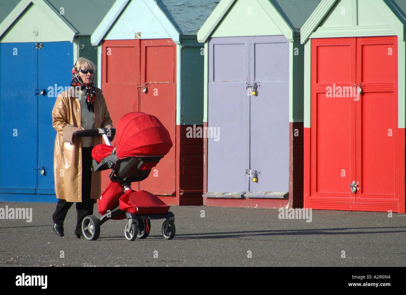 woman pushing a pram past a row of beach huts wearing a raincoat and sunglasses Brighton U.K. Stock Photo