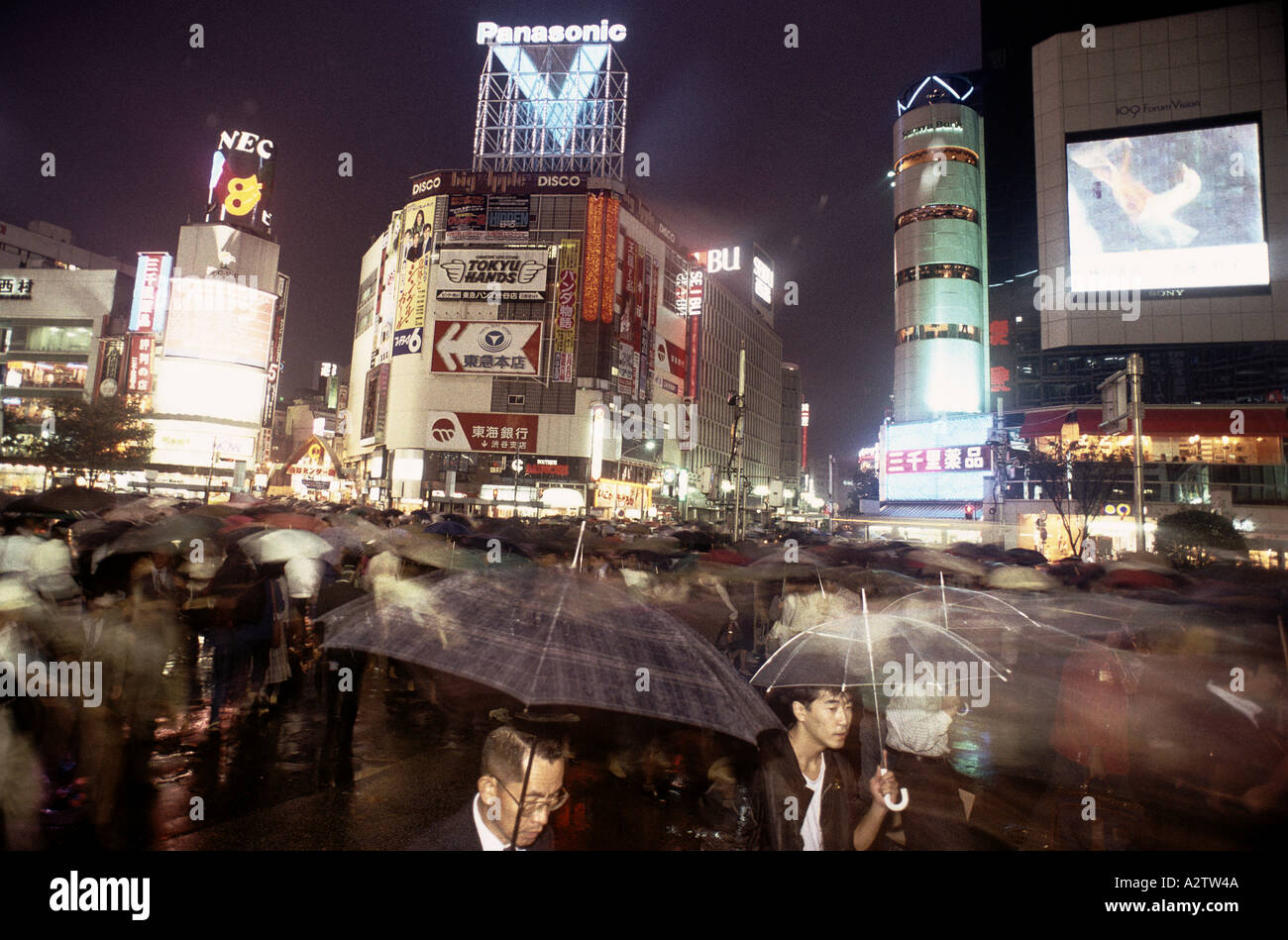 busy street scene at night in the rain in tokyo with tall buildings neon light signs people with umbrellas at cross street Stock Photo