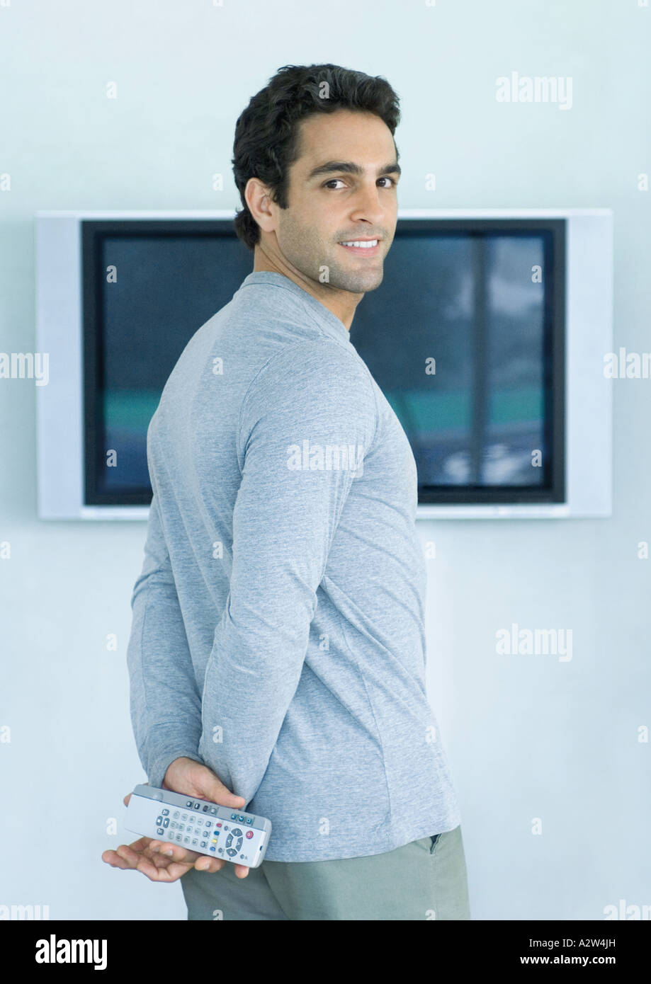 Man standing in front of widescreen TV, holding two remote controls, smiling over shoulder at camera Stock Photo