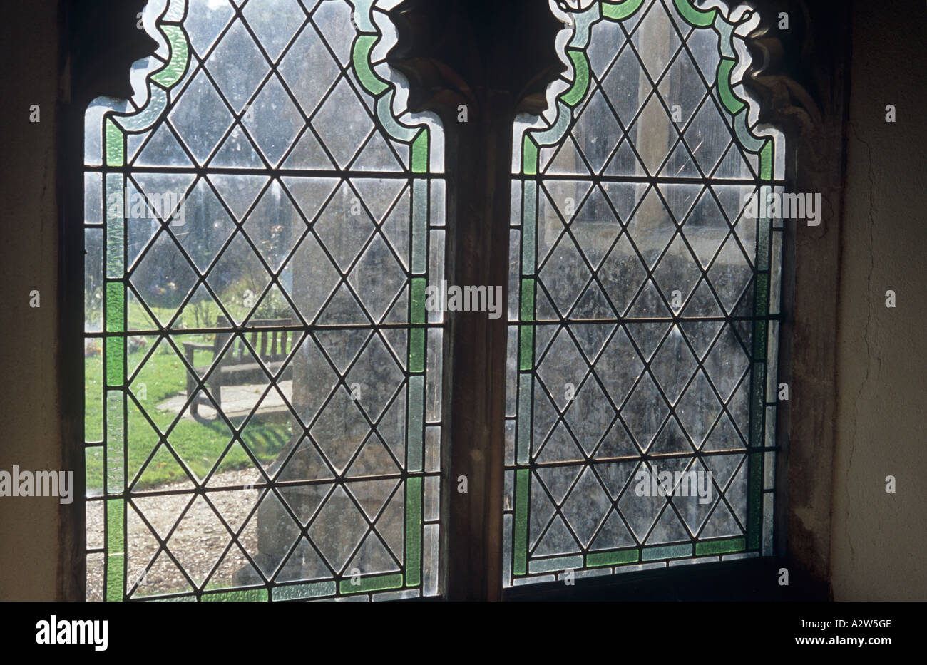 View from inside church through clear diamond leaded windows in Perpendicular style with green fringes to churchyard with bench Stock Photo