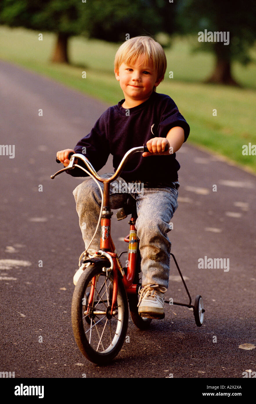 Young fair haired boy on bicycle with stabilizers on road in park. Stock Photo