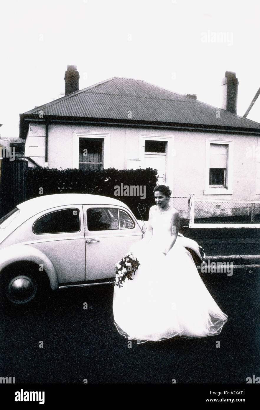Young woman in wedding dress standing by classic Volkswagen Beetle car outside house. Stock Photo