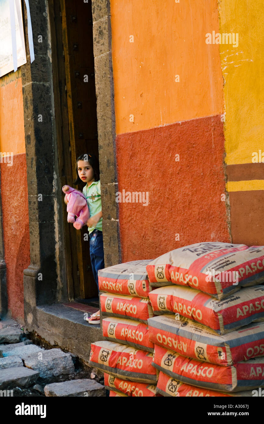 MEXICO San Miguel de Allende Young girl hold doll and stand in doorway of house bags of construction supplies Stock Photo