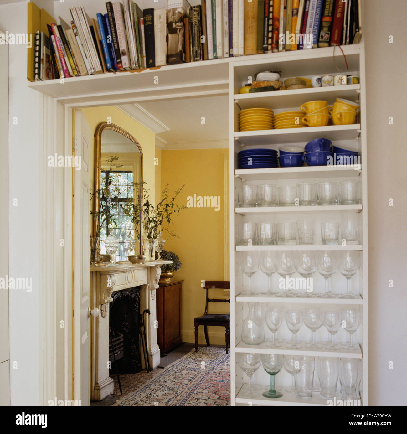Doorway with shelf holding books, crockery and wineglasses looking through to room with mantelpiece. Stock Photo