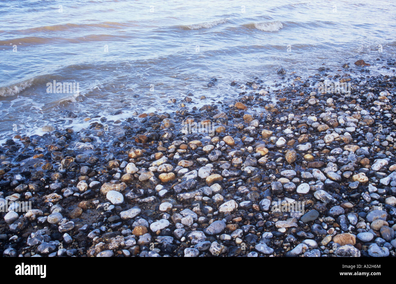 Small waves in a grey sea in warm light breaking onto pebble foreshore Stock Photo