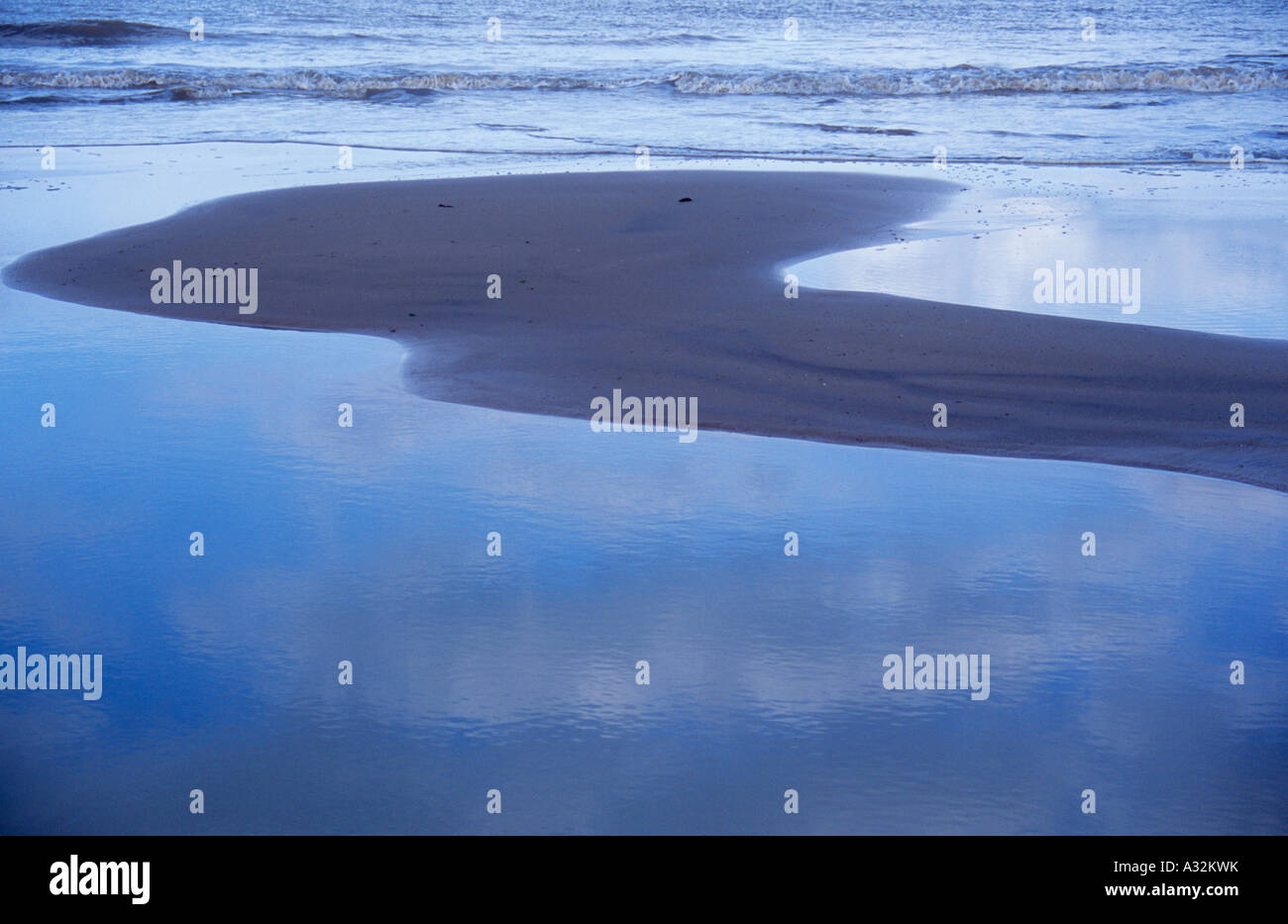 Small waves in a blue-grey sea breaking onto wet sandy beach with only a small dry island of sand and cloud reflections Stock Photo