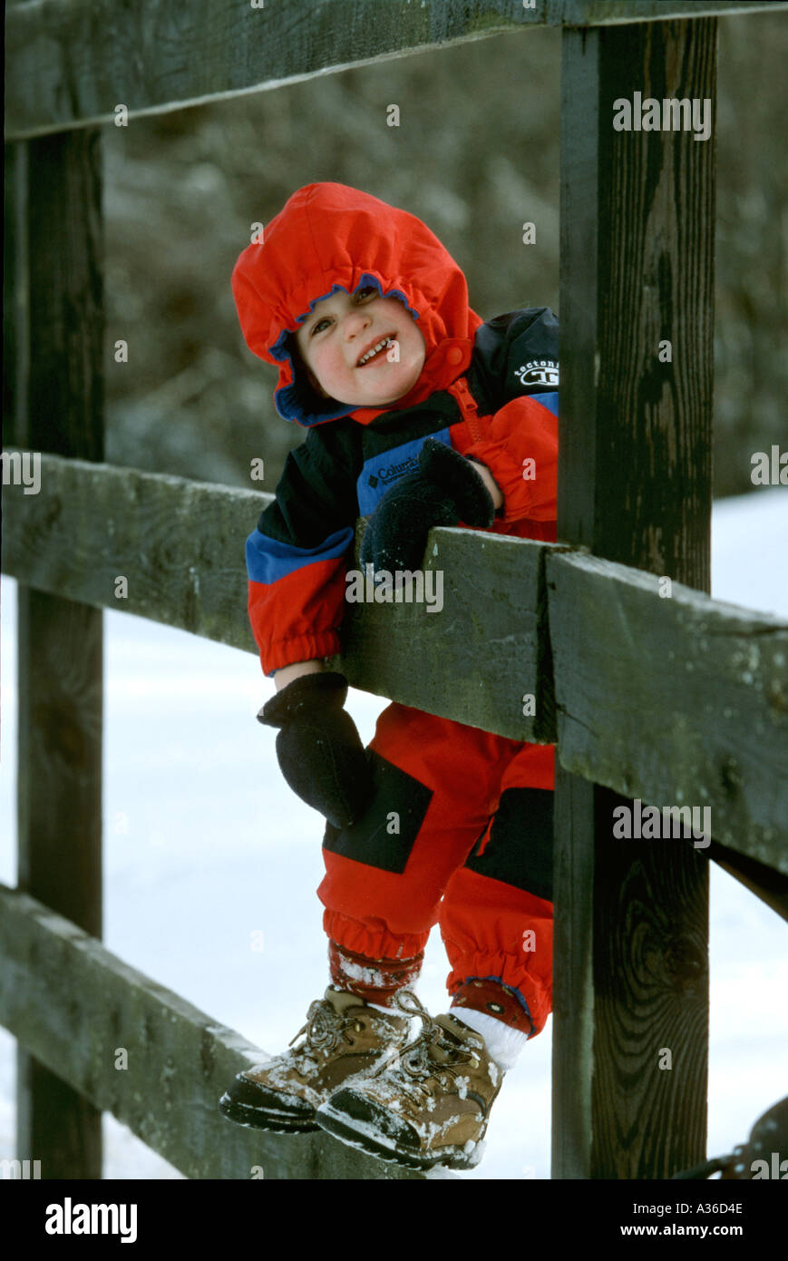 A young girl wearing a bright red ski suit is climbing on a wooden