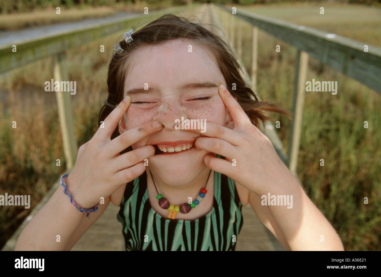 A young girl standing on a boardwalk is looking up at the viewer making a silly goofy face with the help of her fingers Stock Photo