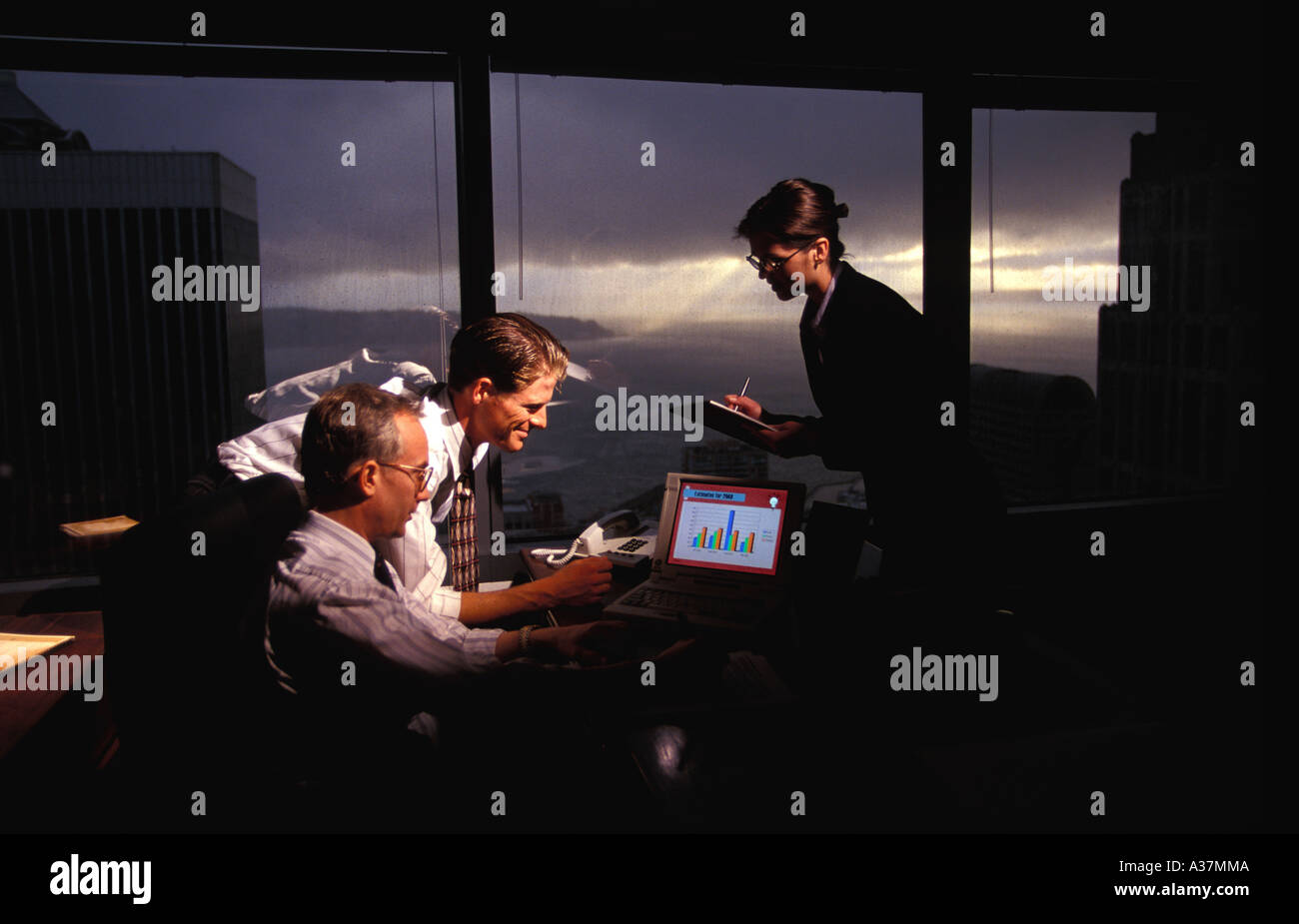 Two executives and a secretary confer in a high rise office while a rain squall storms outside Stock Photo