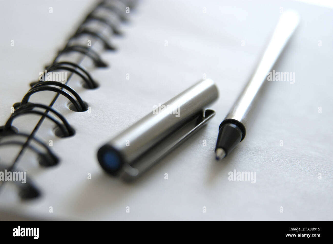 White note book with black spiral binding and stainless steel pen with cap removed. Shot with shallow depth-of-field. Stock Photo