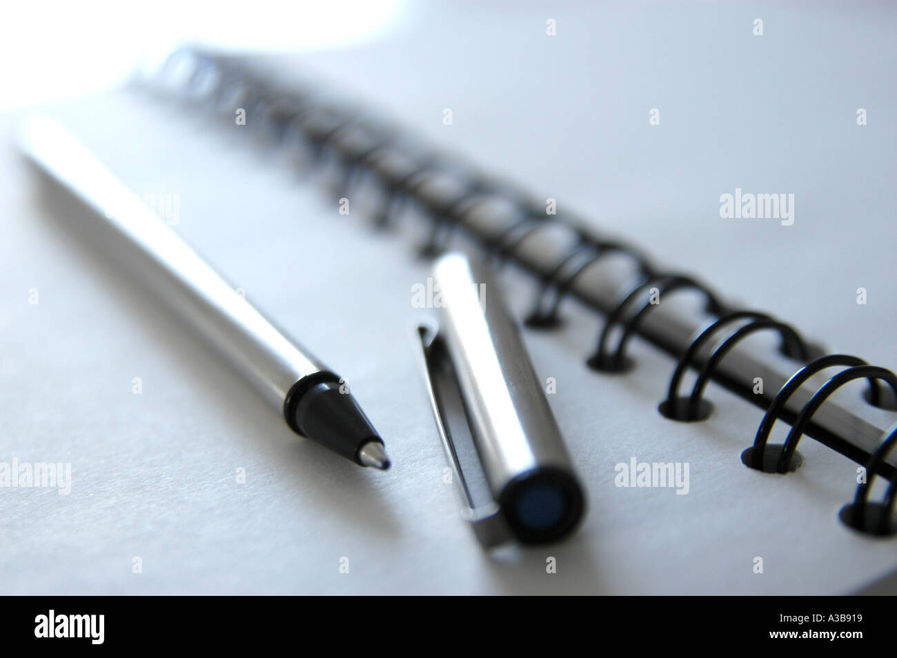 White note book with black spiral binding and stainless steel pen with cap removed. Shot with shallow depth-of-field. Stock Photo