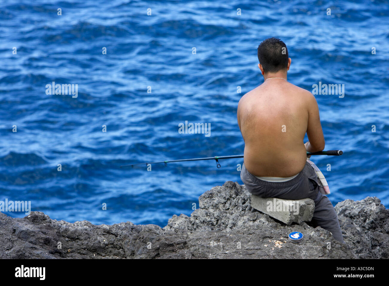 bare backed man sitting on rocks fishing into a very blue sea with rod  Tenerife Canary Islands Spain Stock Photo - Alamy