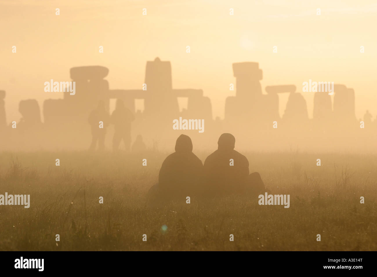 two people sit on the grass watch the sunrise over a misty stonehenge during the summer solstice Stock Photo