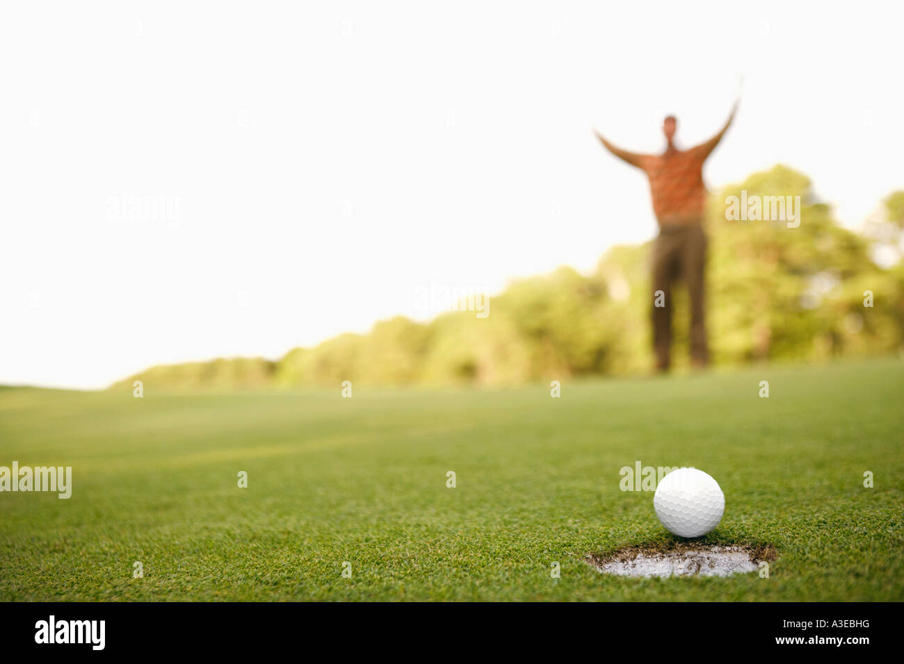 Close-up of a golf ball on the edge of a hole with a man raising his arms in the background Stock Photo